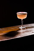 A beautifully presented pisco sour cocktail that sits gracefully on a wooden surface against a dark background. The head is garnished and adds a touch of sophistication and seduction to the scene