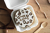 Top view of a stylish bento birthday cake with heart-shaped toppings, presented in a white takeaway box with a wooden spoon next to it on a rustic wooden table.