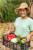 A cheerful woman in a straw hat and glasses holds a box of fresh vegetables, including peppers, aubergines and pumpkins, in a lush garden.