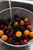 Draufsicht auf frische Brombeeren, Kirschen und goldene Beeren, die in einem Metallsieb unter einem Wasserstrahl gewaschen werden, hervorgehoben durch Wassertröpfchen.