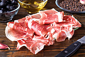 Thinly sliced prosciutto on a wooden board, accompanied by black olives and a bowl of olive oil. A rustic ambience emphasises the gourmet appeal.