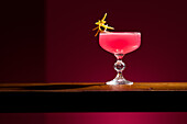 Ein Cosmopolitan-Cocktail wird elegant auf einer hölzernen Oberfläche vor einem dunklen Hintergrund präsentiert. Das leuchtend rosafarbene Getränk ist mit einer Zitrusfrucht garniert und schafft eine anspruchsvolle und einladende Atmosphäre