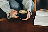 A quiet moment with hands holding a latte in a black mug next to an open book on a wooden table - perfect for depicting relaxation and leisure time