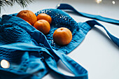 A bright blue string bag filled with juicy mandarins, elegantly placed on a light-coloured table surface with a subtle glow of lights.