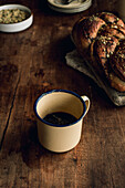 Top view of a freshly baked bakbah bread garnished with pistachios, next to an old tin cup of black coffee, presented on a rustic wooden table.