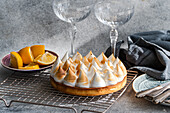 A home-baked lemon cake on a cooling rack next to sliced lemons. Two empty glasses add a touch of elegance to the picture, perfect for a dessert. Soft lighting emphasises the scene.