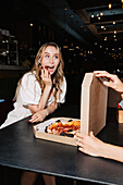 Woman excitedly opens a pizza box at a table with a dimly lit restaurant ambience. Perfect for visual representations of restaurant visits, pizza enjoyment or happy experiences.