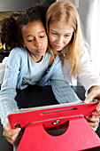 A young woman and a young girl bend over a red tablet, focused and engaged in an interactive activity. The cosy interior environment reflects a warm and modern lifestyle that emphasises learning and connection.