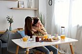 A loving lesbian couple embrace tenderly while eating breakfast at a sunny dining table. The serene setting captures the warmth and intimacy of their relationship