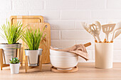 Eine gut organisierte Küchenszene mit hölzernen Schneidebrettern, frischen Grünpflanzen in Töpfen, weißen Rührschüsseln und Utensilien in einem Halter, alles vor einem weißen Backsteinhintergrund.