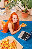 A person enjoys their leisure time on a plush blue carpet with a laptop and a pizza, surrounded by books and a plant The scene represents relaxation and comfort at home