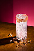 A stunning Paloma cocktail with a salted rim sits on a wooden surface surrounded by ice cubes. The bright pink background emphasises the refreshing look of this classic tequila drink