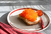 Top view of healthy red organic caviar from trout fish, served on a toasted slice of bread, placed on a white plate with a red border. The place setting includes a red dotted napkin and a spoon.