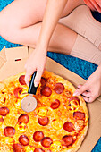 A person in cosy socks sits on a blue carpet and slices a pepperoni pizza in a box with a pizza cutter The surroundings are reminiscent of a warm, relaxed atmosphere at home