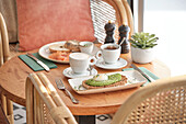 A cosy breakfast scene with avocado toast with poached egg, bagels with lox and two cups of hot coffee, all on a rustic wooden table in a bright setting