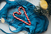 A bright blue drawstring bag lies on a white surface in which two heart-shaped candy canes are gently nestled. Surrounded by soft lights and pine branches, this scene captures a unique holiday mood with a hint of sweetness.