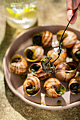 A close-up of an unrecognisable person with a fork eating a dish of baked snails garnished with melted butter and parsley in warm outdoor lighting.