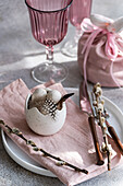 Pastel pink table decoration for Easter. A white ceramic egg holder decorated with a delicate feather is surrounded by pink jars and a matching fabric bag, as well as pussy willow branches on a textured grey background.