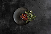 Top view of a dark textured plate filled with shiny Greek grilled olives, garnished with fresh green olive leaves that have a rustic and appetising appearance, ideal for Mediterranean cuisine themes.