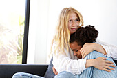 A young woman lovingly embraces a young girl on the couch and provides comfort and security. This warm moment emphasises trust, care and emotional connection in a quiet home environment.