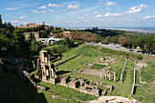 Erkundung der Ruinen eines römischen Theaters in Volterra, Toskana, inmitten einer malerischen Landschaft mit sanften Hügeln und historischer Architektur unter einem strahlend blauen Himmel