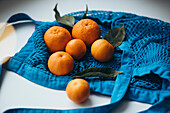 Fresh mandarins with green leaves, arranged in a bright blue string bag on a white table.