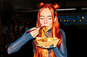A woman with flowing hair savours noodles at an Asian food market and captures the essence of street food.