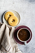 Close-up of a warm coffee cup on a decorative plate paired with golden shortbread biscuits on a small bowl, set on a textured table with a linen napkin for a cosy breakfast atmosphere