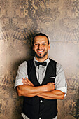 A professional bartender stands confidently with his arms folded, wearing an elegant waistcoat and bow tie. The elegant wallpaper background emphasises the sophisticated atmosphere of a cocktail bar.