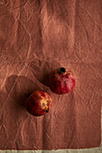 Two ripe pomegranates rest on a textured brown fabric, creating a rustic and earthy still life. The composition highlights natural colours and textures in a simple, elegant scene.
