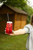 A person with blue painted nails holds a glass of iced red drink with a straw in front of a background of greenery and a wooden structure on a summer day