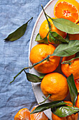 Fresh mandarins with bright green leaves are skilfully arranged on a textured blue fabric, creating a striking contrast. Some of them are peeled to reveal juicy segments.