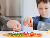 Close-up of sibling hands sprinkling cheese and arranging vegetables on pizza dough. The scene shows creativity and teamwork during a fun cooking activity.