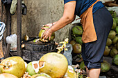 A vendor skilfully peels fresh coconuts with a chopping block at a street market. The scene shows the traditional preparation of coconut water, surrounded by ripe coconuts and tools.