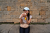 An Asian woman in a blue cap and white shirt enjoys a sandwich while sitting in front of a rustic brick wall in Volterra, Tuscany