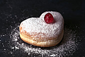 A delicious heart-shaped cream bun, generously sprinkled with icing sugar and decorated with a shiny red cherry on top, served on a dark slate background.