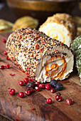 Close-up of a seasoned cheese roll spread with mixed herbs and peppercorns and sprinkled with berries on a rustic wooden table.