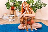 A person with bright red hair enjoys a cosy moment at home, sitting on a blue carpet, hugging a playful dog, surrounded by a pile of colourful books and lush green plants