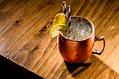 A classic Moscow Mule served in a traditional copper mug on a rustic wooden table. The drink is garnished with a slice of lime, crushed ice and striped straws, creating a refreshing atmosphere