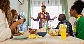 A cheerful birthday party with friends gathered around a dining table. Decorated with party hats and colourful wreaths of flowers, they share drinks, food and laughter.