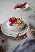 Cut-out, unrecognisable hands of a person serving a delicious redcurrant and passionfruit pavlova on a decorative plate, with the fluffy meringue with bright berries and sliced passionfruit presented against a muted background.