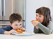 Two young siblings happily eat pizza in a modern kitchen, showing the simplicity of childhood happiness and the pleasure of a universally popular meal.