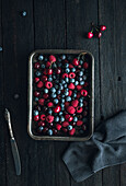 A rustic tray filled with fresh blueberries, raspberries and cherries on a dark wooden background. The bright colours contrast beautifully with the wood and convey a natural, earthy feel.