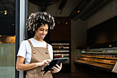 A café employee with vitiligo and an apron works in a modern café with a digital tablet. The atmosphere is cosy and various products are displayed on the shelves