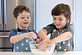 Zwei Kinder in Schürzen mit Sternenmuster mischen Teig in einer Glasschüssel. Sie lächeln und zeigen Teamarbeit in einer modernen Küche, die fröhliche Familienmomente schafft.