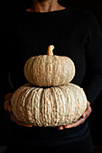 A person holds two stacked pumpkins in front of a dark background. The textured surface of the pumpkins creates an autumnal mood, ideal for seasonal themes and designs.
