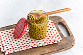 A jar of fresh green, home-made organic peas with a wooden spoon rests on a dotted cloth on a wooden board. The red lid with white polka dots adds a charming, rustic touch.