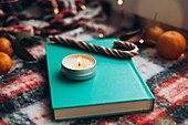 Peaceful Christmas atmosphere with a turquoise book and a burning candle on a colourful, festive blanket, surrounded by a candy cane and mandarins.