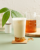 A refreshing iced milk tea with frothy cream, served in a transparent mug with amber jelly resting on the bottom, accompanied by a mug of brewed tea on a wooden coaster