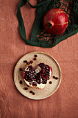 A whole pomegranate and seeds lie on a rustic plate against an earthy background, accompanied by a green mesh bag. The image emphasises the bright colour and texture of the fruit.